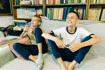 Teenagers play together on the console using joysticks while sitting at home. © Ilona