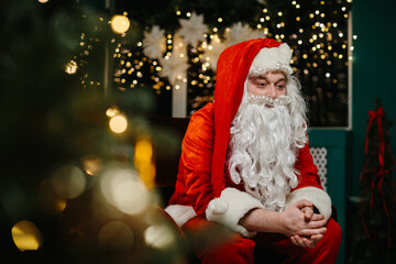 Pensive santa claus sitting near christmas tree with fairy lights