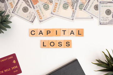 Wooden block tiles with the letters "Capital Loss" on a white background. The word "Capital Loss" is written on a wooden block or wooden cube with money, books, and credit cards on an office desk.
