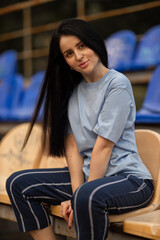 In an empty stadium a brunette girl wearing a blue T shirt sits relaxed on a wooden bench. She appears thoughtful as she enjoys a moment of calm in the sports arena.