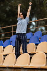 brunette girl wearing blue T shirt stands on empty podium stadium with her hands raised high in celebration expressing pure joy and excitement in moment.