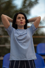 A brunette girl wearing a blue T shirt stands in an empty stadium her hands raised behind her head. She appears shocked and delighted by her unexpected win capturing a moment of excitement and joy.
