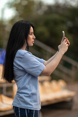 A brunette girl in a blue T shirt stands in the empty stands and focuses on filming a game on her phone. The surroundings are quiet and relaxed highlighting her concentration.