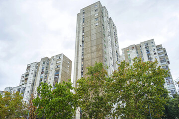 Obraz premium A cityscape from a Yugoslav neighborhood in Belgrade: a bottom-up view of tall concrete apartment buildings against a gray sky. Trees and multi-story houses. Brutalist architecture in Eastern Europe.
