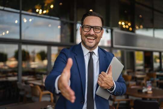 Businessman offering handshake in front of restaurant holding laptop - Powered by Adobe