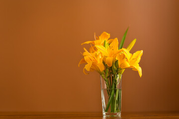 Glass vase filled with blooming orange daylilies on a brown background