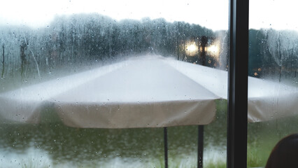 White patio umbrella seen through a rain-covered window, evoking a calm, rainy outdoor atmosphere.