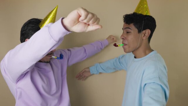 Two young Latinos with golden party hats and party blowers celebrate by jumping with joy and happiness, concept of fun, youthful enthusiasm and festive moment on a light brown background