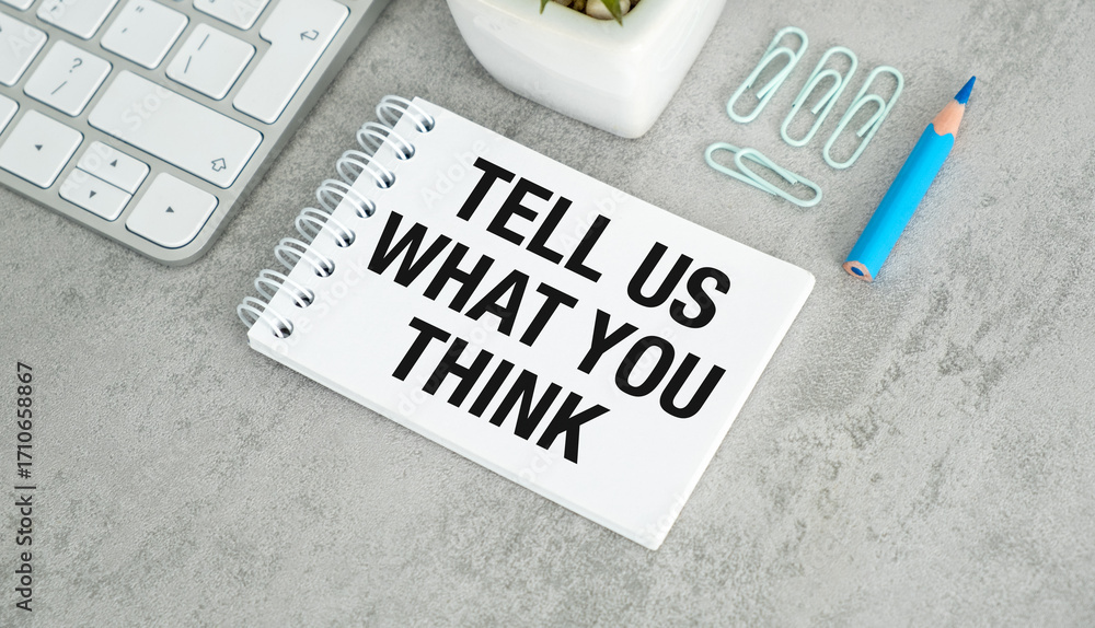Poster tell us what you think writing on a sheet of notebook with pencil, keyboard on a gray table
