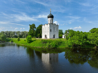 Obraz premium A wide aerial view of the Church of the Intercession on the Nerl, showing the white stone church with its black dome and golden cross reflected in the calm river.