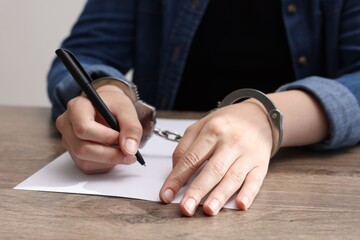 Woman in metal handcuffs writing something at wooden table indoors, closeup