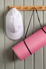 White cap and pink yoga mat hanging on wooden hook rack against gray wall