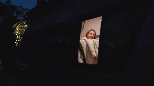 Woman enjoys a cozy night in her van, smiling warmly while wrapped in a blanket and surrounded by soft lights.