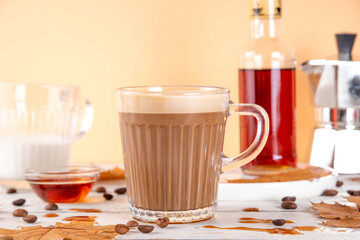 Maple syrup coffee latte drink in glass mug, tasty sweet drink with milky foam, on white table with coffee beans, maple syrup bottle and maple leaves on white wooden table, copy space