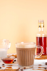 Maple syrup coffee latte drink in glass mug, tasty sweet drink with milky foam, on white table with coffee beans, maple syrup bottle and maple leaves on white wooden table, copy space