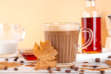 Maple syrup coffee latte drink in glass mug, tasty sweet drink with milky foam, on white table with coffee beans, maple syrup bottle and maple leaves on white wooden table, copy space