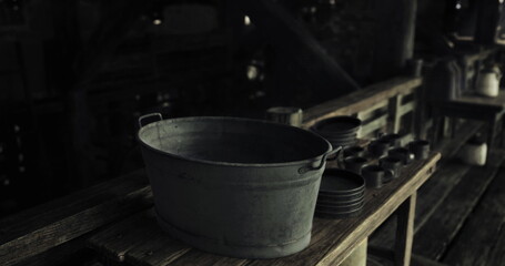 A metal bucket rests on a weathered wooden table surrounded by various dishes in a dimly lit, rustic environment. The scene conveys a sense of history and simplicity.