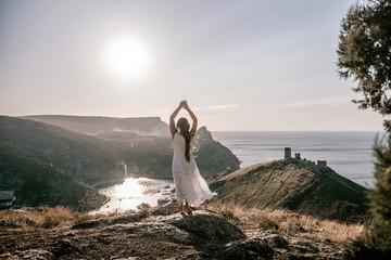 woman stands on a rocky hill overlooking a body of water. She is wearing a white dress and she is...