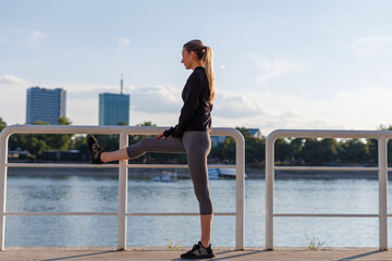 young woman runner stretching her leg by the riverside