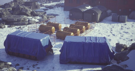 Storage units covered with blue tarps sit in a snowy landscape, surrounded by rocks and yellow tanks. The setting appears to be a remote research location in a cold climate.