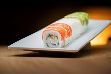 A long sushi roll topped with fresh salmon and avocado slices. The elegant dish, filled with crab and cream cheese, is presented on a modern white plate.