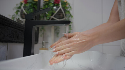 Naklejka premium Woman washing their hands under running water at a sleek black faucet in a bright bathroom setting.