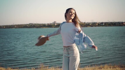 Young woman in shirt, jeans and hat spinning happily by riverside on a sunny windy day. Slow motion cinematic shot, symbolizing joy of life, freedom and travel. 