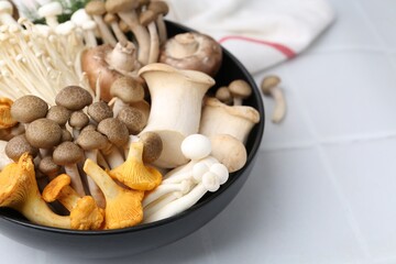 Different raw mushrooms in bowl on white tiled table, closeup. Space for text