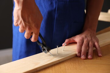 Professional repairman pulling metal nail out of wooden plank with pincers indoors, closeup