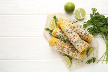 Tasty grilled corncobs with parsley, cheese and lime slices on white wooden table, flat lay. Space for text