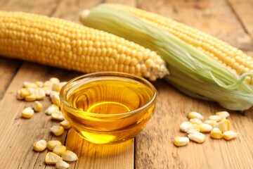 Corn oil in glass bowl, kernels and cobs on wooden table, closeup