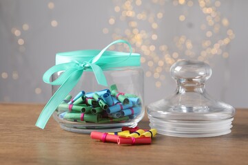 Colorful paper notes in glass jar on wooden table against grey background with blurred lights, closeup. Bokeh effect
