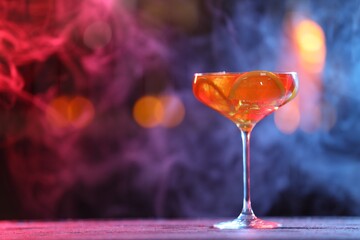 Glass of alcoholic cocktail on table against blurred background with smoke, space for text. Bokeh effect