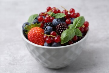Different ripe berries and basil leaves in bowl on light grey textured table, closeup