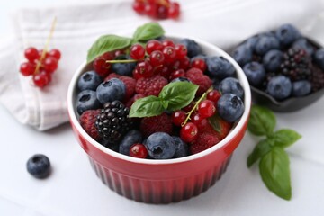 Different ripe berries and basil leaves in bowl on white tiled table, closeup
