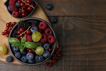 Different ripe juicy berries and basil in bowl on wooden table, flat lay. Space for text