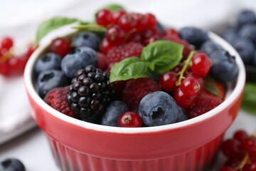 Different ripe berries and basil leaves in bowl on light table, closeup
