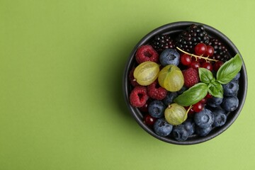 Different ripe juicy berries and basil in bowl on green background, top view. Space for text