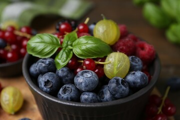 Different ripe juicy berries and basil in bowl on wooden table, closeup