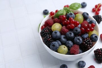 Different ripe juicy berries and basil in bowl on white tiled table, closeup. Space for text