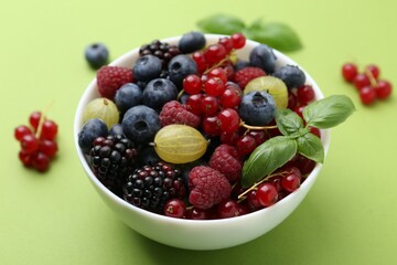 Different ripe juicy berries and basil in bowl on green background, closeup