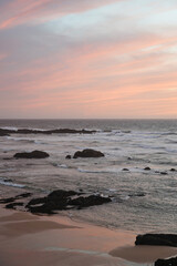 Rocky beach on the coast of Portugal. Waves, foam, pastel sky with pink and purple hues, peaceful scene, natural depth, sunset atmosphere.