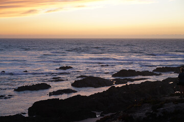 Rocky beach on the coast of Portugal. Waves, foam, pastel sky with yellow and pink hues, peaceful scene, natural depth, sunset atmosphere.