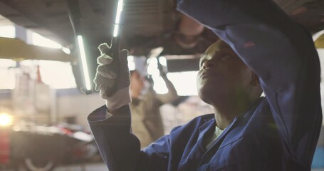 Inspecting Black mechanic in blue coveralls holding LED light under chassis garage bay, copy space