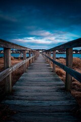 A long wooden walkway stretches towards serene waters, surrounded by tall grass and a twilight sky. The distant town adds a peaceful charm to the scene during the evening