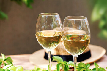 White wine glasses in bright sunlight with straw hat and fresh greenery on wooden table..