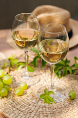 Two glasses of white wine with fresh grapes and straw hat on sunlit rustic table with green foliage..