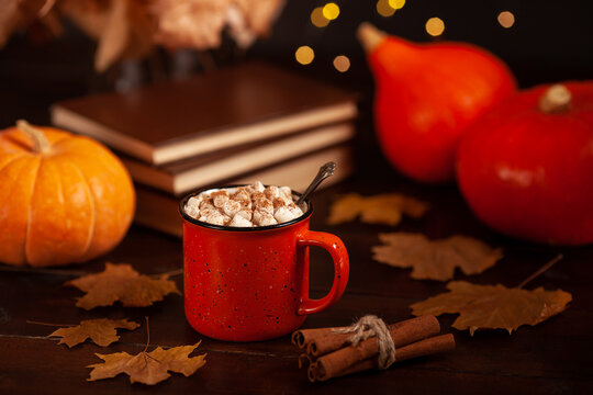 Warm autumn composition with hot chocolate and marshmallows in a red mug, pumpkins, cinnamon sticks, and dry leaves on a rustic wooden table with cozy seasonal atmosphere.