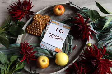 Calendar for October 10: the number 10 and the month's name "October" in English on a flip calendar among dahlias, cookies, and fruit