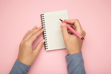 Female's hand writing in white blank open spiral notebook with pen on pastel pink background. Flat lay, copy space.